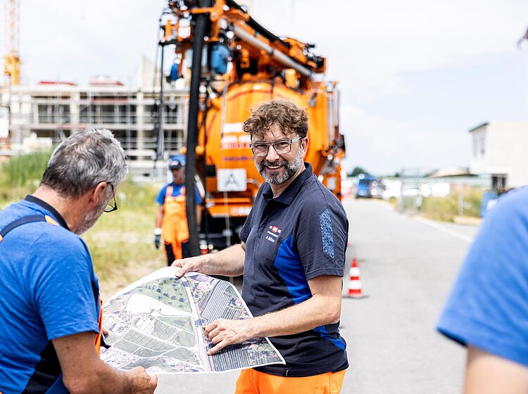 PAKO-Team bei Fahrzeugarbeit Mehrere PAKO-Mitarbeiter in blauer Arbeitskleidung arbeiten gemeinsam an einem orangefarbenen Kanalreinigungsfahrzeug auf dem Betriebshof.