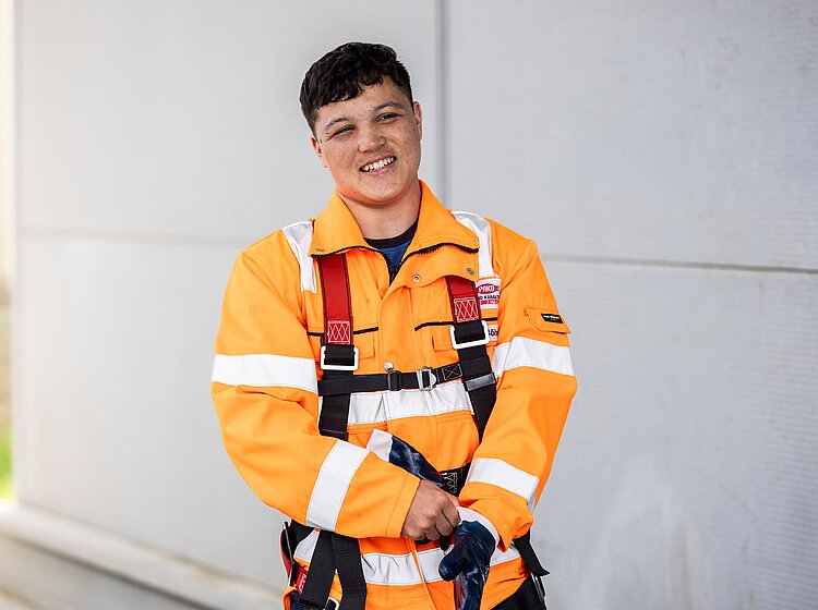 Mitarbeiter in Schutzausrüstung PAKO-Mitarbeiter trägt orange Schutzkleidung mit Reflexstreifen und Sicherheitsgeschirr, steht vor grauer Wand.