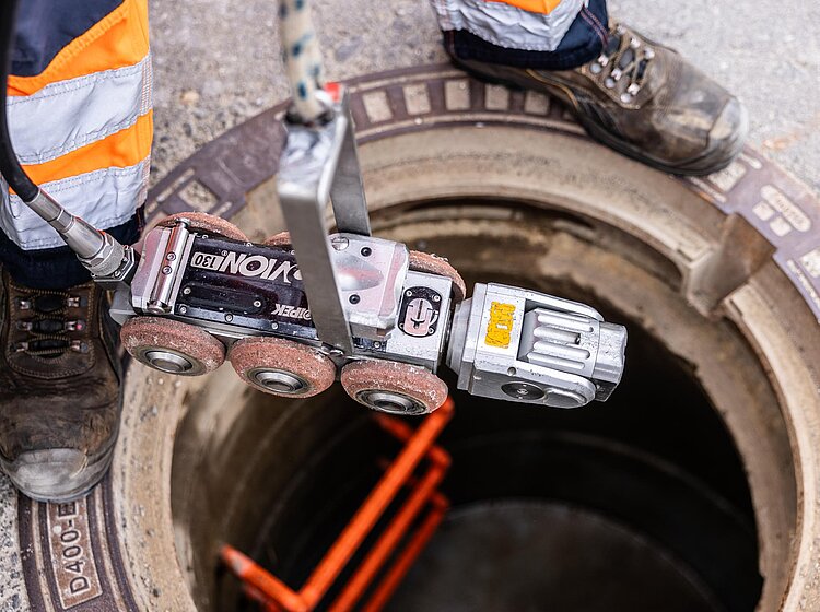 Kamera in Rohrleitung einsetzen Blick in einen Schacht, in dem eine Fahrwagenkamera mit orangefarbenem Kabel in eine Rohrleitung eingeführt wird.