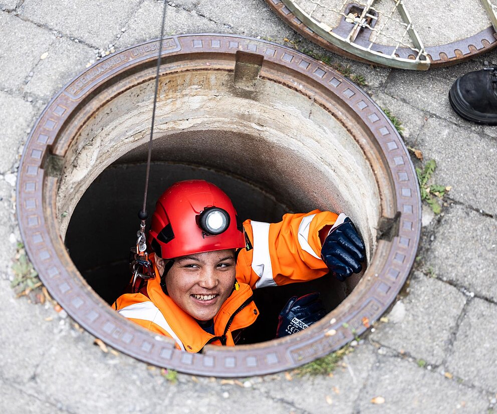 Inspektion im Kanalschacht PAKO-Mitarbeiter mit Helm und Schutzkleidung schaut aus einem geöffneten Kanalschacht, umgeben von Pflastersteinen.
