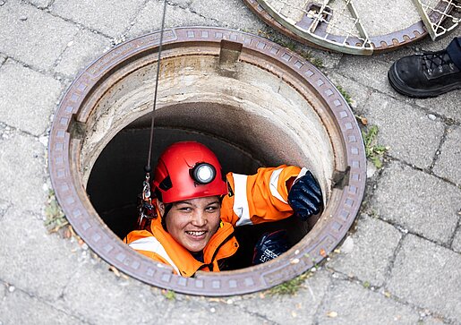 Inspektion im Kanalschacht PAKO-Mitarbeiter mit Helm und Schutzkleidung schaut aus einem geöffneten Kanalschacht, umgeben von Pflastersteinen.