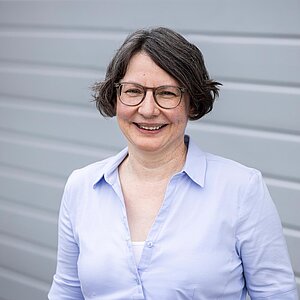 Birgit Böhm Mitarbeiterin der PAKO Rohr- und Kanaltechnik GmbH & Co. KG mit heller Bluse, lächelt in die Kamera vor einer grauen Wand.