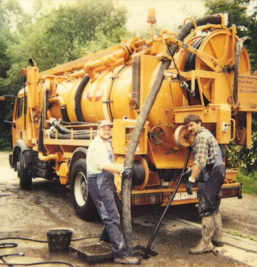 Arbeiten am Kanalfahrzeug Zwei Mitarbeiter der PAKO Rohr- und Kanaltechnik GmbH & Co. KG arbeiten an einem großen orangefarbenen Kanalfahrzeug auf einer Baustelle.