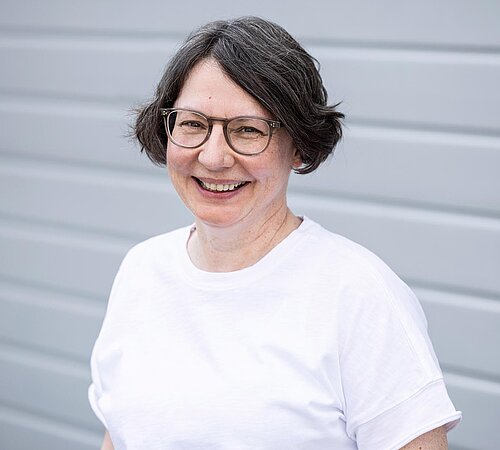 Birgit Böhm Mitarbeiterin der PAKO Rohr- und Kanaltechnik GmbH & Co. KG mit Brille, weißem T-Shirt, steht vor einer grauen Wand und lächelt.