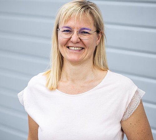 Portrait Sandra Prophet Mitarbeiterin der PAKO Rohr- und Kanaltechnik GmbH & Co. KG steht lächelnd vor einer grauen Wand, trägt ein weißes Oberteil.