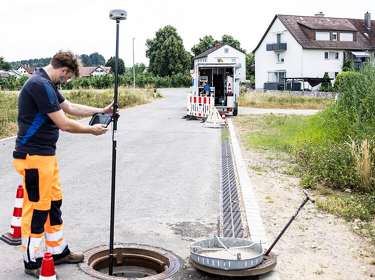 Vermessung von Kontrollschacht PAKO-Mitarbeiter misst einen geöffneten Kontrollschacht mit Messgerät auf einer Straße, Absperrung vorhanden.