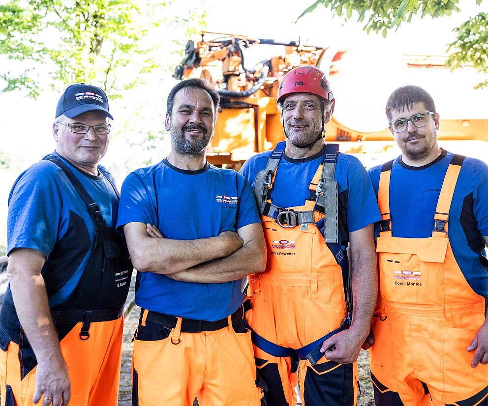 PAKO-Team im Freien Fünf PAKO-Mitarbeiter in blauer Arbeitskleidung und orangenen Arbeitshosen stehen lachend zusammen im Freien.