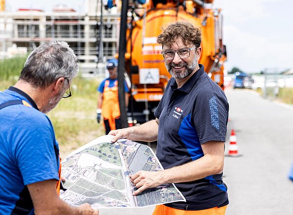 Teamarbeit am Einsatzfahrzeug PAKO-Mitarbeiter besprechen den Arbeitseinsatz neben einem orangefarbenen Kanalreinigungsfahrzeug, einer hält einen Ordner in der Hand.