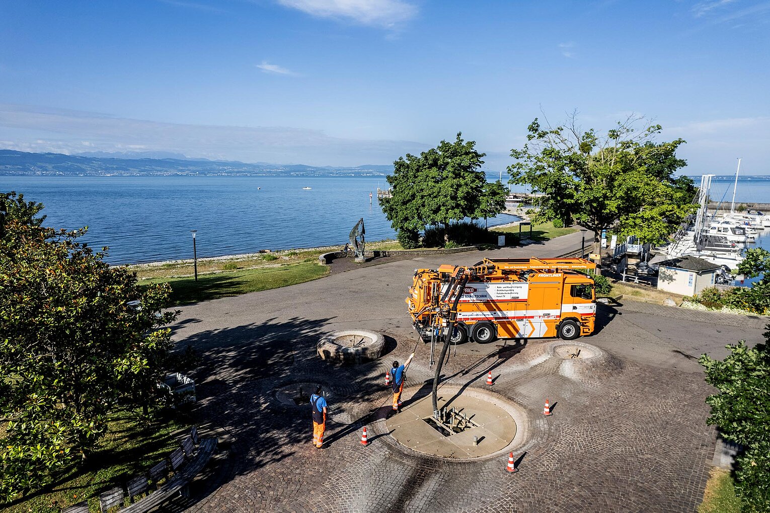 Straßenbaustelle mit Seeblick PAKO-Fahrzeuge und Absperrungen auf einer Straßenbaustelle, im Hintergrund Blick auf einen See und grüne Landschaft.