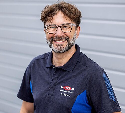Alexander Böhm Mitarbeiter der PAKO Rohr- und Kanaltechnik GmbH & Co. KG mit Bart, Brille und dunkelblauem T-Shirt, steht vor einer grauen Wand.