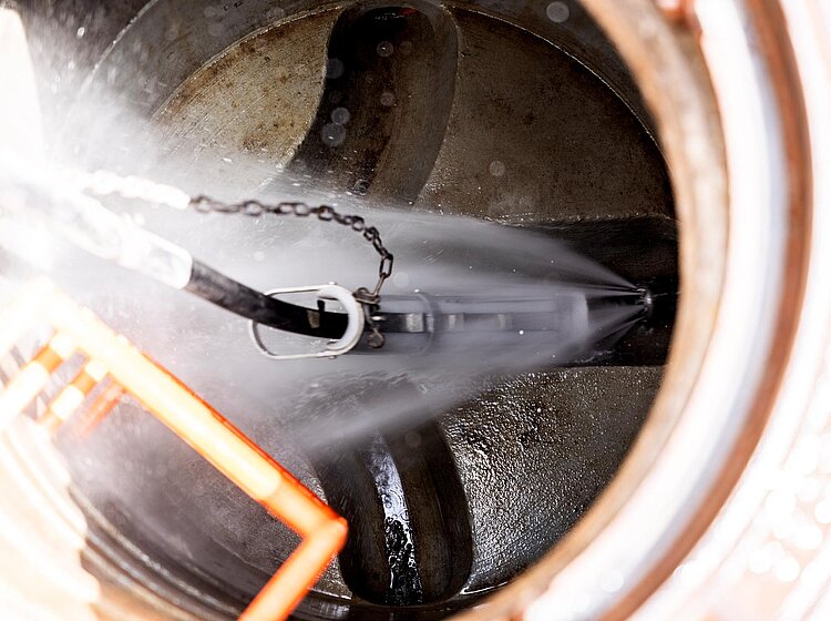 Kanalreinigung durch Schachtdeckel Blick in einen offenen Schacht mit Reinigungsgerät, das im Kanalrohr arbeitet; Details der Rohrreinigung sichtbar.