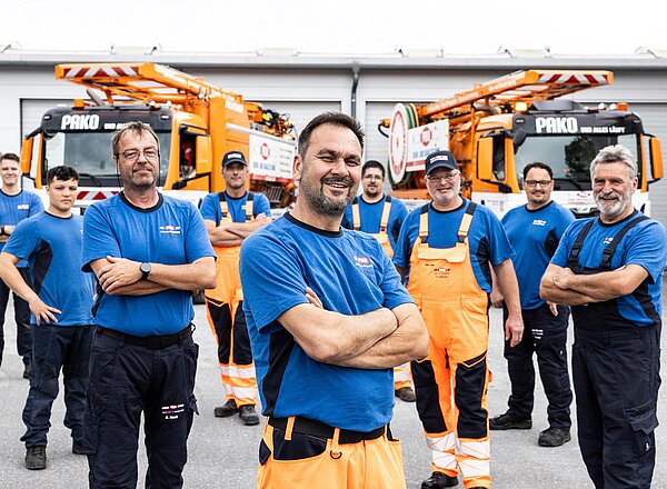 Gruppenfoto PAKO-Team Mehrere PAKO-Mitarbeiter in blauer Arbeitskleidung stehen Arm in Arm nebeneinander und lächeln in die Kamera.