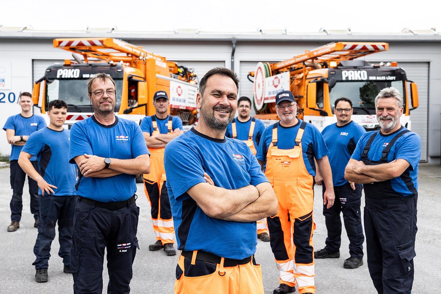 Gruppenfoto PAKO-Team Mehrere PAKO-Mitarbeiter in blauer Arbeitskleidung stehen Arm in Arm nebeneinander und lächeln in die Kamera.