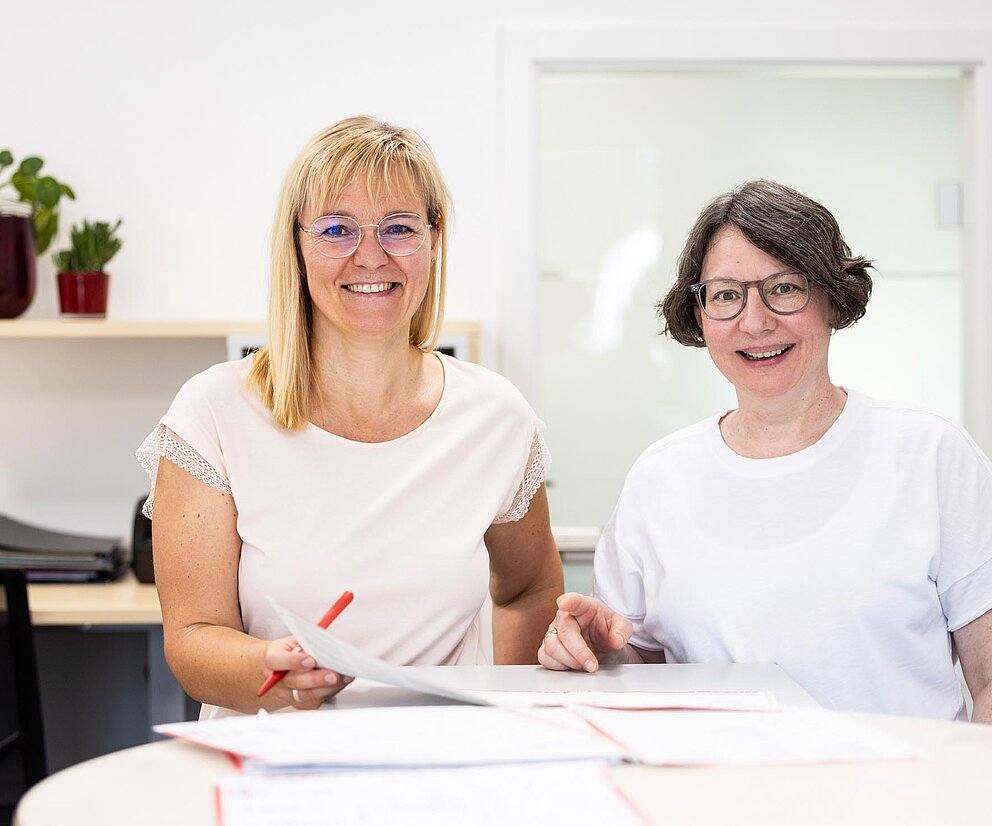 Empfangsteam im Büro Zwei Mitarbeiterinnen der PAKO Rohr- und Kanaltechnik GmbH & Co. KG sitzen am Empfangstresen und lächeln in die Kamera.
